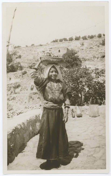 Black-and-white photo of a young woman carrying a large basket filled with items on her head. She stands on a stone road in front of a barren hill.