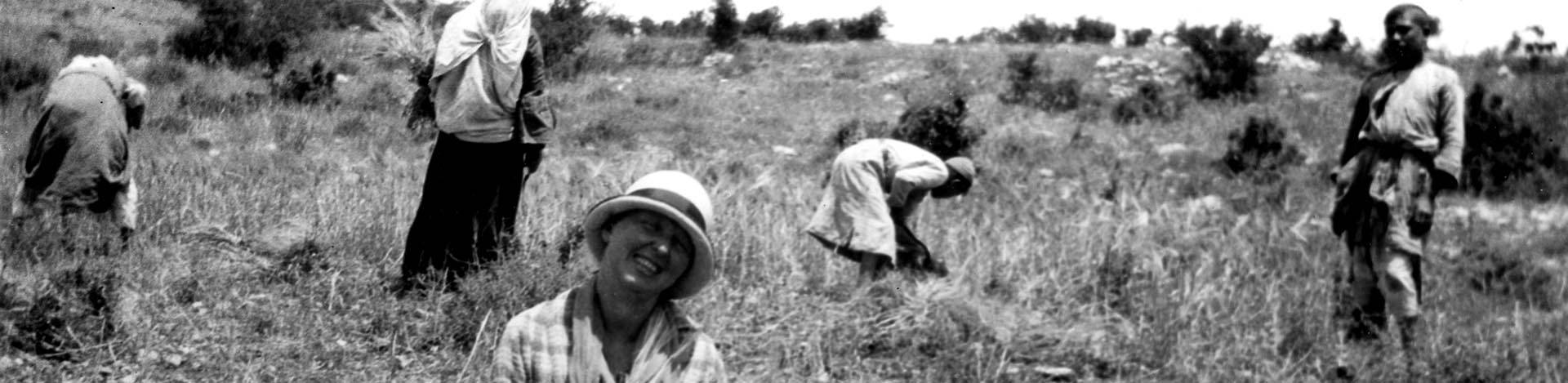 Black and white photo of five people working outdoors in a meadow. Four dark-skinned people in the background are bent over with their backs to the camera. A light-skinned woman in a hat in the foreground is smiling at the camera.